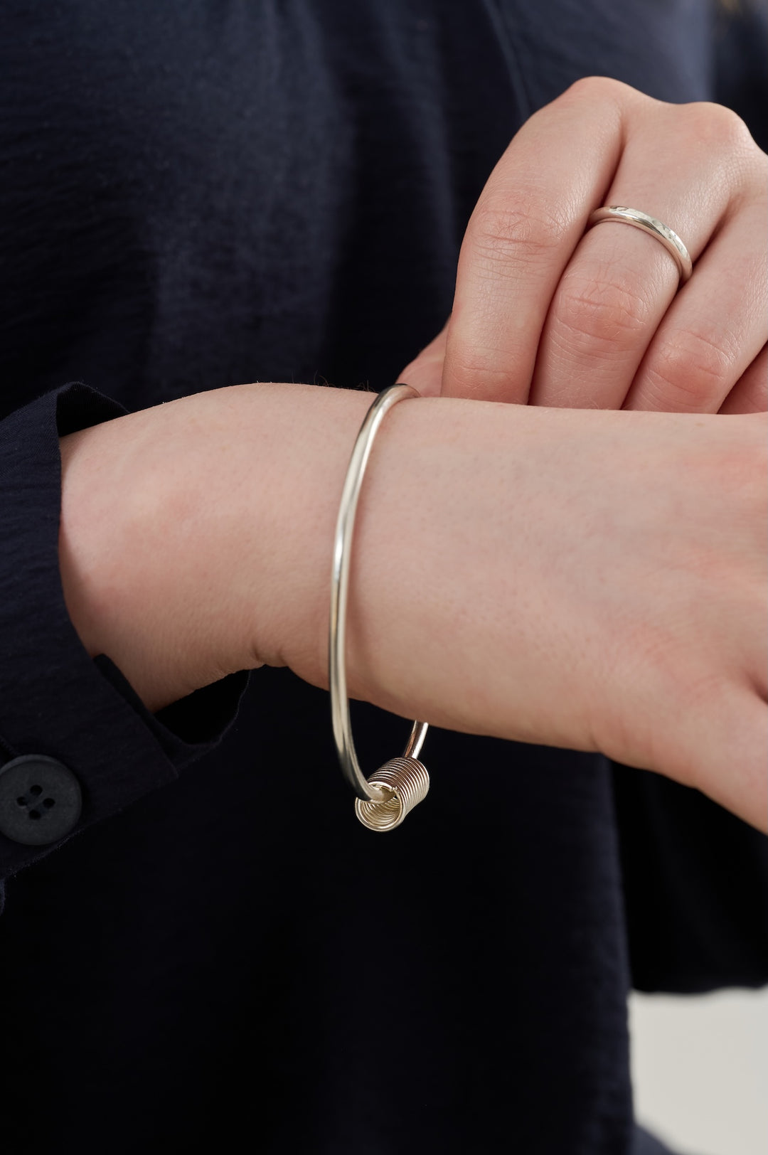 Sterling silver bangle made from 3mm wide silver wire with a 16x8mm silver coil hanging on it in foreground and textured stacker ring in background